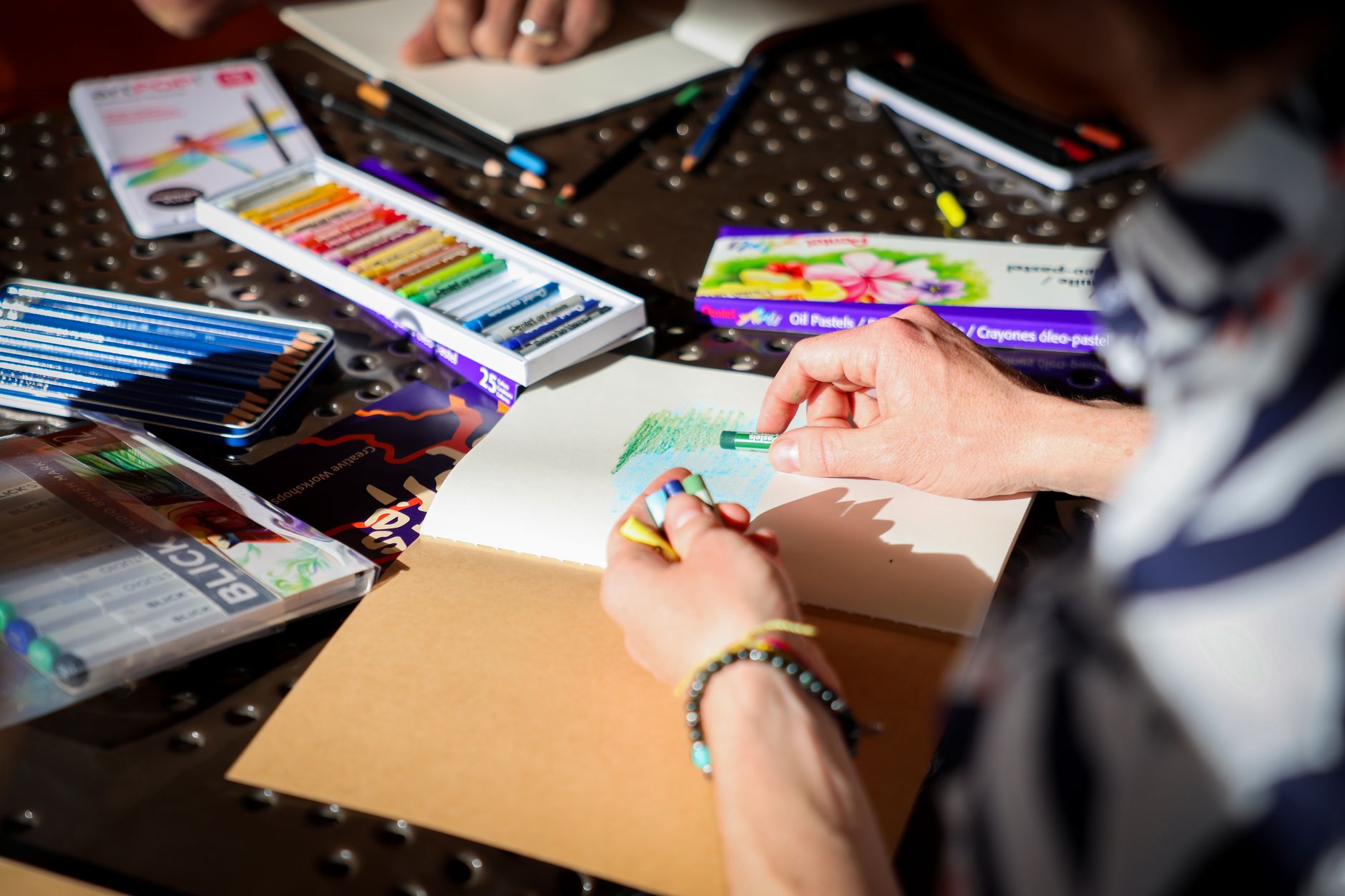 Hands color a drawing on paper with crayons, surrounded by art supplies on a table.